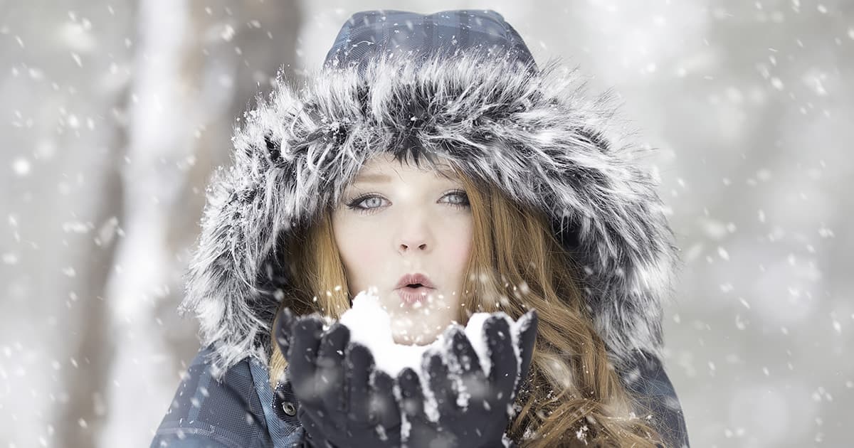 A young woman bundled up in the cold, blowing snow from her hands.