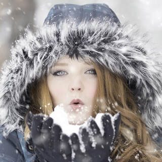 A young woman bundled up in the cold, blowing snow from her hands.